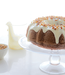 Hummingbird Bundt Cake on cake stand with dripping frosting and pecans.