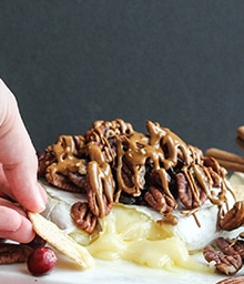 Person dipping cracker in Caramel, Pecan, and Cranberry Baked Brie.