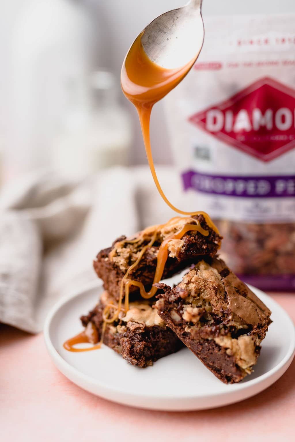 Spoon drizzling caramel on plate of Salted Caramel Pecan Brookies in front of bag of Diamond nuts.